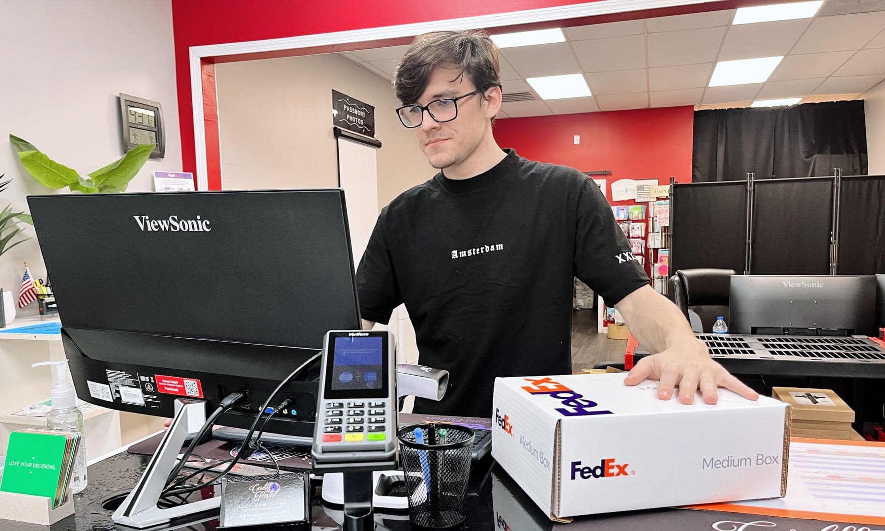 A Cardiff Postal team member at the front counter beside a register and a FedEx box, the red accent wall behind