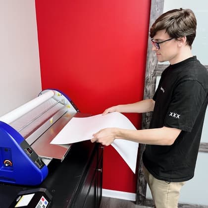 A Cardiff Postal team member feeding a large print through a blue laminating machine in front of the red wall