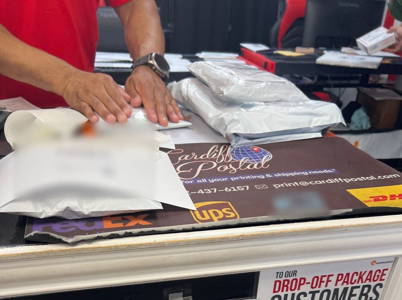 Hands pressing a mailer flat at the counter, more mailers stacked beside (labels blurred)
